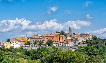 Vibrant hillside village with colorful buildings, historic tower, lush greenery, under blue sky with clouds, showcasing picturesque architecture and scenic beauty in Italy.
