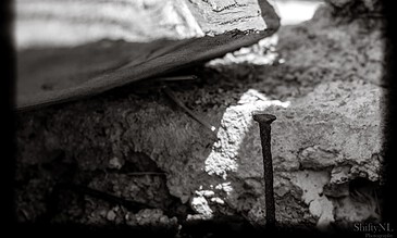 Rusted nail embedded in dirt beneath weathered wooden plank, black and white close-up photography for vintage or abandoned site visuals.