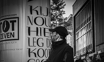 A candid street shot capturing daily life with focus on a man in motion among city surroundings. A man wearing a cap and jacket walks past a large advertisement billboard in an urban setting, with pedestrians and modern buildings in the background.