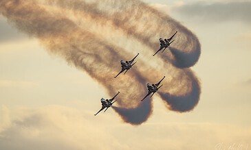 Spectacular image of four fighter jets performing an aerial maneuver, creating smoke trails in the sky at sunset, showcasing precision and power. Aero combat jets flying in formation, leaving smoke trails against a cloudy sky during sunset.
