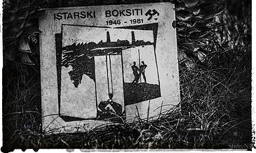 A memorial plaque for Istarski Boksiti, featuring a black and white illustration of two figures, with dates 1946-1981, surrounded by grass and natural elements.