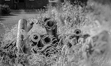 A black and white photo of discarded pipes and debris on the ground, with a building and vegetation in the background.