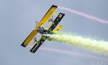 B. An eye-catching yellow biplane executing an aerobatic maneuver, leaving vibrant green and pink smoke trails in the sky during an airshow. A. Vintage yellow biplane performing an aerobatic stunt with colorful smoke trails against a cloudy sky.