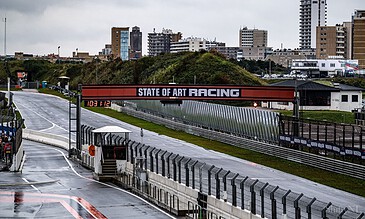 1. Overcast race track with "STATE OF ART RACING" banner and city skyline background on a rainy day, showcasing modern motorsport infrastructure.