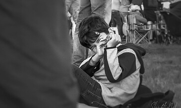 A young boy with glasses and a striped hoodie, sitting on the grass and taking a photograph with a camera during an outdoor event, with people and tables visible in the background. A young boy sitting outdoors, holding a camera up to his face, surrounded by people and outdoor furniture, capturing a moment at an event or gathering.
