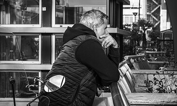 A contemplative man with gray hair in a black puffer vest and hoodie, standing at an outdoor city café, deep in thought near large windows and urban scenery in black and white.