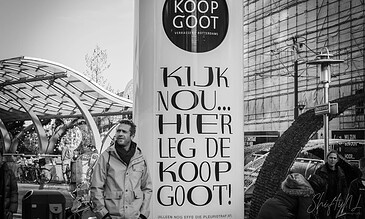 An outdoor urban scene in Rotterdam, featuring a large promotional booth and people walking, with modern architecture and festive decorations in black and white photography.