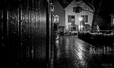 Rainy night scene in a charming European village with illuminated windows, wet cobblestone street, and festive string lights, captured in black and white for a timeless, atmospheric mood.
