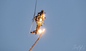 An intense image of a military helicopter performing a rescue maneuver, with flames and smoke trailing as it ascends into the sky. Military helicopter in mid-air during a dramatic rescue operation with bright flames and smoke against a clear blue sky.