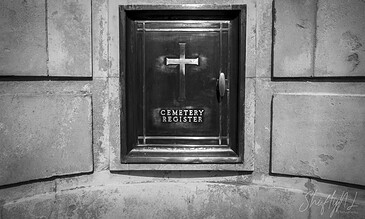 A black and white photograph of a cemetery register box embedded in a stone wall, featuring a cross symbol and the words "Cemetery Register," capturing a somber, timeless atmosphere. A dark, metal cemetery register box with a cross symbol mounted on a stone wall, used for recording burial details in a historic or religious cemetery setting.