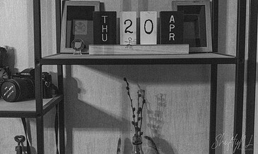 Black and white indoor scene featuring a shelf with a calendar displaying "Thu 20 Apr," a camera, and decorative items, capturing a minimalist and artistic home decor setup.