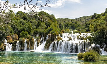 A lush waterfall cascading over rocks into a clear turquoise pool surrounded by green foliage and trees under a partly cloudy sky.