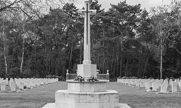 A black and white photograph of a war memorial with a cross at the center, surrounded by rows of headstones in a tranquil cemetery setting. War memorial with a cross and surrounding headstones in a peaceful cemetery, black and white photography.