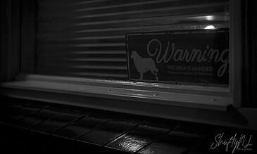 Black and white photo of a warning sign with a dog silhouette behind a window with blinds, indicating a guarded area.