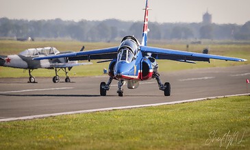 A vibrant jet aircraft is positioned on the runway, ready for departure, captured in sharp detail with a scenic background of the airfield and distant landscape. Colorful jet fighter on runway preparing for takeoff at an airfield, showcasing aviation photography skills.