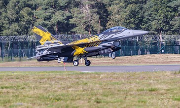 A sleek F-16 fighter jet with striking yellow and black camouflage design, preparing for takeoff on a runway surrounded by greenery. F-16 Fighting Falcon jet taking off on runway with trees in background, showcasing military aviation and speed.