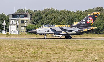 A military jet with tiger artwork on its tail, parked on the runway with a control tower in the background, captured during a clear day. F-16 Fighting Falcon jet aircraft on runway with tiger-themed tail design, near control tower, during daytime.