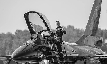Close-up of a pilot standing on a fighter jet, ready for flight, with a clear sky and distant trees in the background. Aero pilot preparing a fighter jet for takeoff on a clear day.