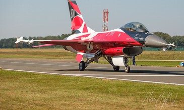 Close-up of an F-16 fighter jet parked on the runway, highlighting its aerodynamic shape and striking color scheme against a bright sky. F-16 fighter jet on runway with clear sky, showcasing sleek design and vibrant red, black, and gray colors.