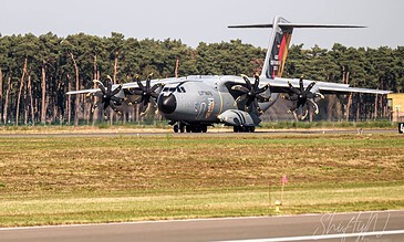 Close-up of a military cargo plane on the tarmac, showcasing its four propeller engines and tail design, captured during an airshow or aircraft event. Aero L-100 military transport aircraft parked on runway with forest background.