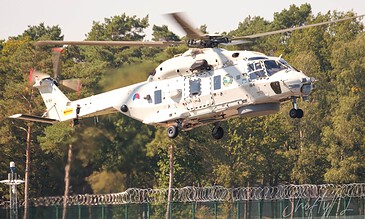 A white helicopter flying low over a secure fenced area, with lush green trees in the background, showcasing aviation photography and dynamic motion. Aerial view of a white helicopter in flight above a fenced area with trees in the background, capturing a dynamic moment during a flight demonstration or training exercise.
