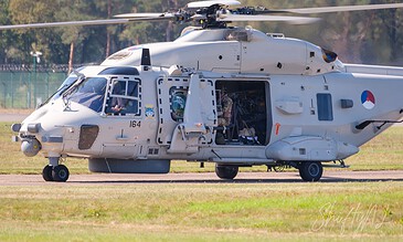Close-up of a military helicopter on the runway, showcasing its detailed design and open side door, set against a background of trees and clear sky. Aerial view of a white military helicopter on the runway with open side door, ready for takeoff or landing.