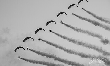 Aerial display of six paragliders flying in formation, leaving trails of smoke in the sky during a daytime event.
