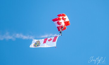 Aerial view of a skydiver descending with a Canadian flag and a red parachute against a clear blue sky, showcasing dynamic motion and vibrant colors.