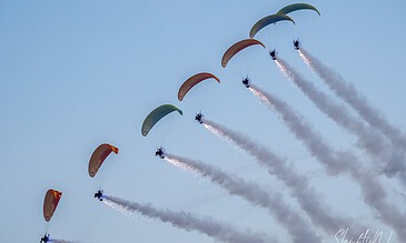 Aerial display of six colorful paragliders flying in formation against a clear blue sky.