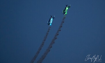 A flying display of two illuminated aircraft performing an aerobatic maneuver against a dark blue sky, leaving smoke trails behind them.