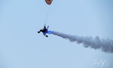 Aero paraglider soaring through the clear blue sky, leaving a trail of white smoke behind during a flight demonstration.