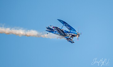Aerial photograph of a blue aerobatic aircraft performing a stunt in clear blue sky, leaving white smoke trail behind.