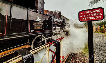 A historic steam train emitting white steam, parked near a red and black warning sign that reads "De toegang tot den spoorweg is verboden" (Access to the railway is prohibited), with trees in the background.