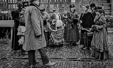 Vintage dressed group of women and men participating in a historical reenactment outdoors, wearing elaborate period costumes with accessories, on a cobblestone street with old buildings in the background.