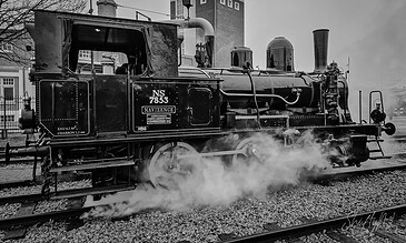 Steam-powered vintage train on railway tracks with smoke and historic buildings in the background, captured in black and white.