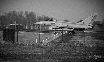 A sleek fighter jet sits on the runway behind a fence, highlighting military aircraft readiness and defense systems, all captured in a striking black-and-white photograph. Fighter jet parked on runway behind a fence, capturing a powerful military aviation scene in black and white; showcases aircraft readiness and defense technology.