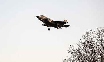 A modern F-35 stealth fighter jet soaring in the sky, capturing a dramatic shot with a minimalist background and natural elements like tree branches for contrast. F-35 fighter jet in flight against clear sky with tree branches in the foreground, showcasing advanced military aircraft features and sleek design.
