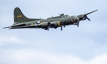 Vintage B-25 Mitchell aircraft in flight, painted in military camouflage with distinctive nose art and checkered pattern, soaring through a cloudy sky.