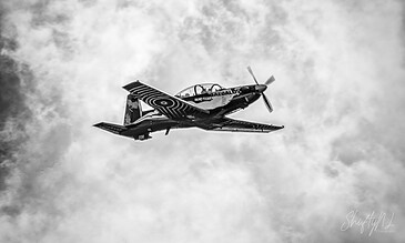 A black and white photograph of a training plane flying through a cloudy sky, showcasing dynamic motion and classic aircraft design.