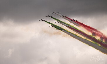 Vibrant Italian Air Force aerobatic team flying in formation with colorful smoke trails against a cloudy sky.