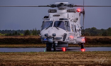 RN-03 military helicopter starting up on a wet airstrip at dusk with crew members visible inside, illuminated by red and blue lights, capturing a dramatic moment of flight operation.