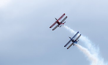 Red and blue aerobatic planes performing mid-air stunt with smoke trails against a cloudy sky.