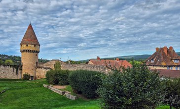 Historic European village featuring a medieval stone tower, lush greenery, and classic architecture with red-tiled roofs. Medieval stone tower with red-tiled conical roof and fortified walls in a scenic European village under a partly cloudy sky.