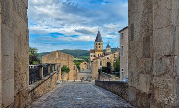 Charming view of a historic European village with cobblestone streets, stone buildings, and prominent church steeples against scenic countryside backdrop. Historic European village street with cobblestone path and medieval church towers under blue skies.
