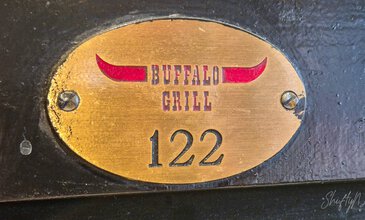 Close-up of a brass plaque engraved with "Buffalo Grill" featuring red horn graphics and the number 122, attached to a black textured surface, showcasing craftsmanship and branding of a high-quality outdoor grill. Glossy brass plaque with "Buffalo Grill" logo featuring red horns and the number 122, mounted on a dark textured surface, part of a professional barbecue grill setup.