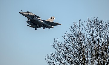 French fighter jet flying low over trees against clear blue sky, capturing a dynamic military aircraft in flight.