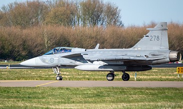 Military Saab Grippen fighter jet taxiing on runway during daytime with autumn trees in background.