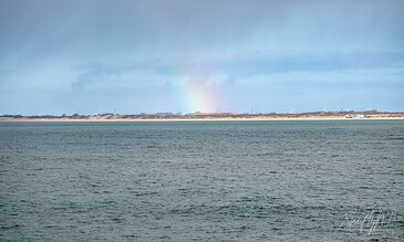 Capturing the serene beauty of nature, this image showcases a vibrant rainbow arching across the sky above tranquil waters. A stunning view of a rainbow over a calm ocean with a distant shoreline under a partly cloudy sky.