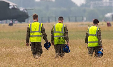 Three 845 Naval Air Squadron crew members in hi-vis vests walking through the grass with their helmets during the APROC 2018 personnel recovery exercise at Gilze-Rijen Air Base, the Netherlands, June 2018.