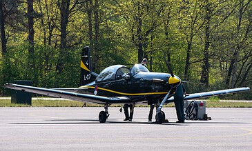 Black Pilatus PC-7 trainer with yellow details parked on the grass at Woensdrecht Air Base, the Netherlands, during a visit to 131 EMVO Squadron, May 2017.